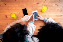 Сверху неузнаваемые многонациональные молодые гомосексуальные мужчины просматривают социальные сети на смартфоне и пьют свежие напитки, улыбаясь, сидя за столиком кафе во время романтического свидания — стоковое фото