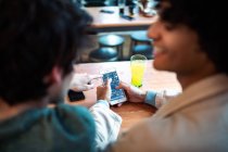 Back view of unrecognizable multiethnic young homosexual men browsing social media on smartphone and having fresh drinks smiling while sitting at cafe table during romantic date — Stock Photo
