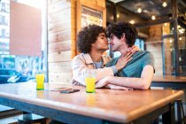 Vue latérale d'hommes ethniques excités s'embrassant avec les yeux fermés sur la table et riant pendant un rendez-vous romantique dans la cafétéria moderne — Photo de stock
