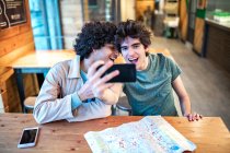 Мультиэтнические молодые гомосексуальные мужчины делают селфи на смартфоне и пьют свежие напитки, улыбаясь, сидя за столиком кафе во время романтического свидания — стоковое фото