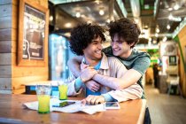 Багатоетнічні молоді гомосексуальні чоловіки обіймаються, дивлячись на навігаційну карту напрямку і маючи свіжі напої, посміхаючись, сидячи за столом кафе під час романтичного побачення — стокове фото