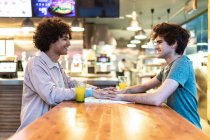 Seitenansicht aufgeregter ethnischer Männer, die sich umarmen, die Hände über dem Tisch halten und beim romantischen Date in der modernen Cafeteria lachen — Stockfoto