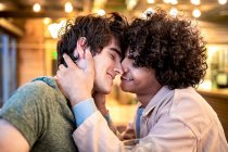 Seitenansicht aufgeregter ethnischer Männer, die sich mit geschlossenen Augen am Tisch umarmen und beim romantischen Date in der modernen Cafeteria lachen — Stockfoto