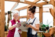 Adult woman writing on clipboard near elderly colleague while working in hothouse together — Stock Photo