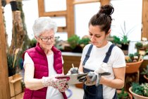 Adult woman writing on clipboard near elderly colleague while working in hothouse together — Stock Photo