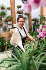Vista laterale del giovane giardiniere che sorride e annaffia fiori e piante in fiore durante il lavoro in aranciera di legno — Foto stock