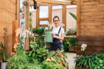 Glückliche erwachsene Frau lächelt in die Kamera und trägt Dose mit blühender Blume bei der Arbeit im Terrassengarten — Stockfoto
