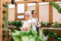 Vista lateral de mujeres ancianas y adultas positivas con guantes y gafas sonriendo para la cámara y cruzando brazos mientras trabajan juntos en el jardín interior - foto de stock