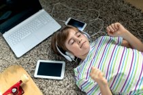 Von oben ein fröhlicher kleiner Junge in lässigem Hemd, der mit Kopfhörern Musik hört, während er auf dem Teppich neben Geräten und Skateboard im Schlafzimmer liegt — Stockfoto