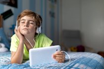 Positive preteen boy in casual wear with headphones lying on bed and watching movie on tablet while resting at home — Stock Photo