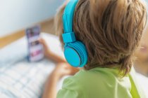 Visão traseira menino em desgaste casual com fones de ouvido deitado na cama e assistindo filme no smartphone enquanto descansa em casa — Fotografia de Stock