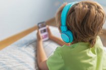 Хлопчик спиною в повсякденному одязі з навушниками лежить на ліжку і дивиться фільм на смартфон під час відпочинку вдома — стокове фото