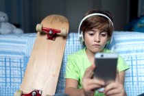 Niño pequeño con auriculares escuchando música y charlando con amigos en la red social mientras está sentado cerca de monopatín en el dormitorio - foto de stock