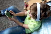 Маленький хлопчик з навушниками слухає музику і спілкується з друзями в соціальній мережі, сидячи біля скейтборду в спальні — стокове фото