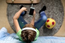 Bambino con le cuffie che ascolta musica e chatta con gli amici sui social network mentre siede vicino allo skateboard in camera da letto — Foto stock