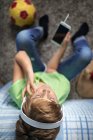 Маленький мальчик с наушниками, слушающий музыку и общающийся с друзьями в социальной сети, сидя рядом со скейтбордом в спальне — стоковое фото