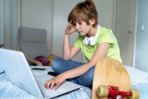 Depressiver kleiner Junge mit Kopfhörern am Hals sitzt auf dem Bett und benutzt Laptop, während er Zeit in der Selbstisolierung wegen des Coronavirus zu Hause verbringt — Stockfoto