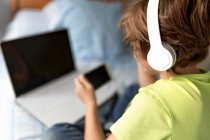 Вид школьника в повседневной одежде и наушниках, играющего в видеоигры на смартфоне дома — стоковое фото