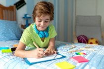 Side view of positive schoolboy in casual wear and wireless headphones enjoying music and drawing with pencils while spending free time in bedroom — Stock Photo