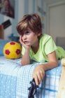 Скучный маленький мальчик в повседневной одежде лежит на кровати рядом с мячом и скейтбордом недовольный самоизоляцией дома — стоковое фото