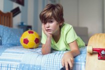 Скучный маленький мальчик в повседневной одежде лежит на кровати рядом с мячом и скейтбордом недовольный самоизоляцией дома — стоковое фото