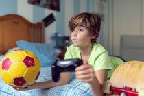 Серйозний маленький хлопчик проводить час вдома і грає в відеоігри, лежачи на ліжку з м'ячем і скейтбордом, розміщеним поруч — стокове фото
