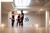 Jeune homme et femmes avec des nattes parlant entre eux tout en se tenant dans un studio spacieux pendant la formation de yoga — Photo de stock