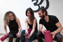 Joyeux jeune femme souriant et montrant les données sur smartphone aux amis enchantés avec des nattes avant l'entraînement de yoga en studio — Photo de stock