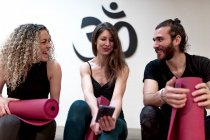 Mujer joven feliz sonriendo y mostrando datos en el teléfono inteligente a amigos encantados con esteras antes del entrenamiento de yoga en el estudio - foto de stock