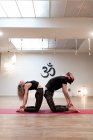 Vue latérale du jeune homme et de la jeune femme en vêtements de sport se pencher en arrière et toucher les pieds tout en faisant l'exercice Ustrasana pendant l'entraînement de yoga dans un studio spacieux — Photo de stock