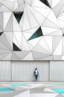 Бородатий хлопець дивиться спосіб, тримаючи йогу під рукою, спираючись на металеву стіну в сучасній будівлі з восьмикутною стелею — стокове фото