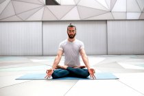 Бородатый парень в спортивной одежде сидит в позе лотоса и медитирует с закрытыми глазами и жестом мудры в просторной комнате с геометрической стеной и полом — стоковое фото