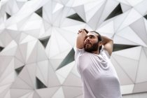Снизу бородатый парень в белой футболке смотрит в сторону и делает упражнения на растяжку для рук во время занятий йогой против декоративной геометрической стены — стоковое фото