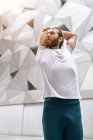 From below bearded guy in white t shirt looking away and doing stretching exercise for arms while doing yoga training against ornamental geometric wall — Stock Photo