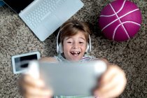 De cima de menino alegre em fones de ouvido deitado no chão perto de gadgets e bola e jogar videogame em tablet enquanto passa o tempo em casa — Fotografia de Stock