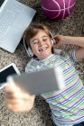 Зверху веселий маленький хлопчик у повсякденній сорочці слухає музику з навушниками, лежачи на килимі біля гаджетів і скейтборду в спальні — стокове фото