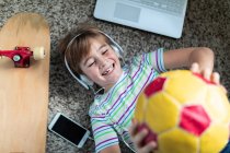 Von oben ein fröhlicher Junge in Freizeitkleidung, der mit Kopfhörern und Smartphone Musik hört und Ball wirft, während er im Zimmer auf Teppich liegt — Stockfoto