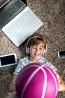 Сверху веселый мальчик в повседневной одежде слушает музыку с наушниками и смартфоном и бросает мяч, лежа на ковре в комнате — стоковое фото