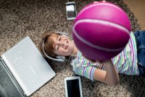 Сверху веселый мальчик в повседневной одежде слушает музыку с наушниками и смартфоном и бросает мяч, лежа на ковре в комнате — стоковое фото