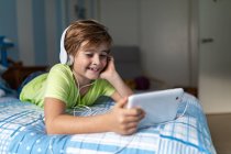 Positive preteen boy in casual wear with headphones lying on bed and watching movie on tablet while resting at home — Stock Photo