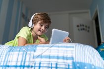 Positiver Frühchen-Junge in Freizeitkleidung mit Kopfhörern auf dem Bett liegend und Film auf Tablet anschauend, während er zu Hause ruht — Stockfoto