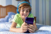 Positiver Frühchen-Junge in Freizeitkleidung mit Kopfhörern, die auf dem Bett liegen und während er zu Hause ruht, Filme auf dem Smartphone ansehen — Stockfoto