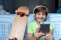 Смеющийся мальчик в наушниках, слушающий музыку и общающийся с друзьями в социальной сети, сидя рядом со скейтбордом в спальне — стоковое фото