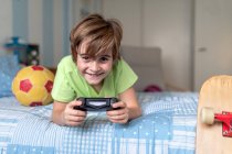 Cheerful little boy spending time at home and playing video game while lying on bed with ball and skateboard placed nearby — Stock Photo