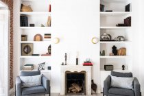 Cómodos asientos grises ubicados cerca de la chimenea y estantería en la acogedora sala de estar en un elegante apartamento - foto de stock