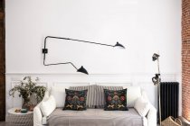 Удобный диван с мягкими подушками расположен под современной металлической лампой возле столов с украшениями в стильной уютной гостиной на дому — стоковое фото