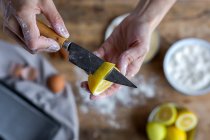 From above crop hand of unrecognizable woman covered in flour peeling lemon and showing to the camera a fresh half cut lemon with knife — Stock Photo