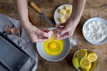 From above top view unrecognizable female breaking fresh chicken egg into bowl while cooking pastry in a wooden table with fresh ingredients — Stock Photo