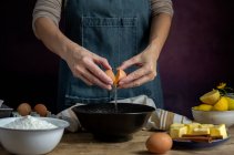 Невизначена жінка розбиває свіже куряче яйце в миску, готуючи випічку в дерев'яному столі зі свіжими інгредієнтами — стокове фото