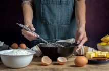 Coltiva anonima donna in grembiule montando uova in ciotola nera su tavolo di legno con limone, farina, burro e bastoncini di cannella ingredienti per la torta — Foto stock